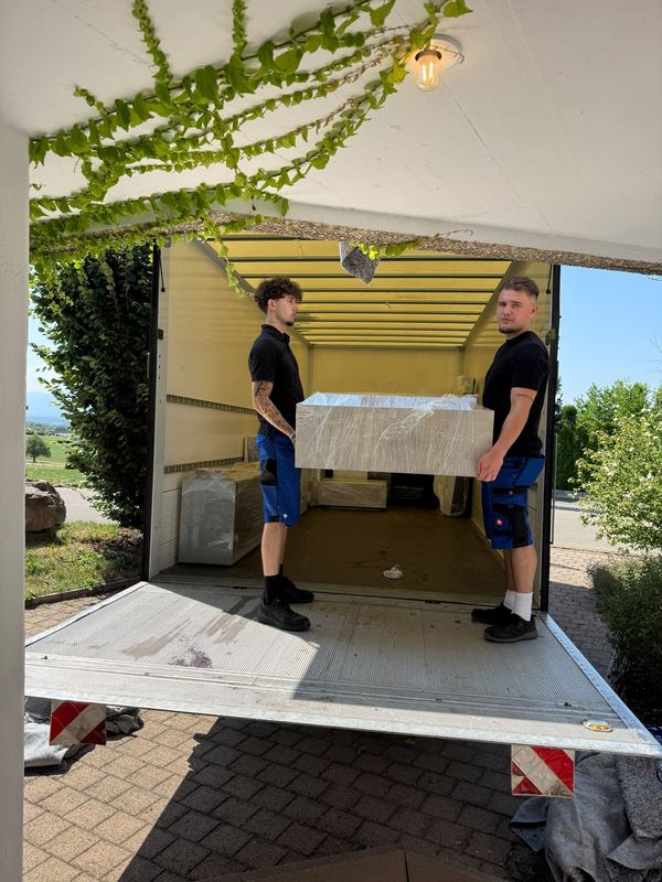 Verpacktes Möbelstück in den Wagen heben Zwei Mitarbeiter hieven ein folienverpacktes Möbelstück über die abgesenkte Ladebordwand hinein in den Laderaum des Umzugswagens