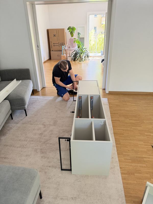 Aufbau des Sideboards am Zielort Ein kniender Mitarbeiter baut in einem hellen Wohnzimmer ein weißes Sideboard zusammen, dahinter stehen Umzugskartons
